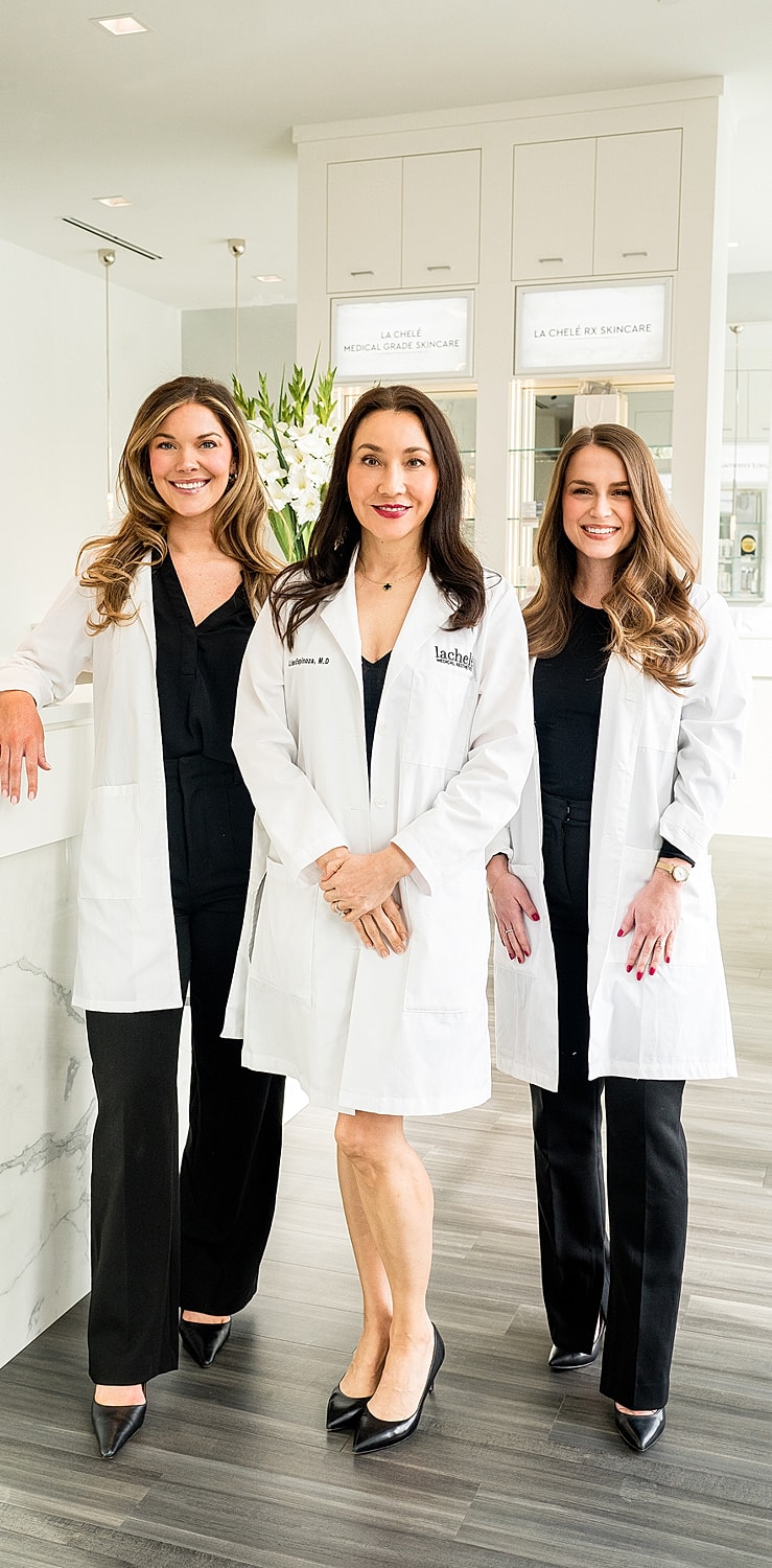 Three women in white lab coats smiling indoors.