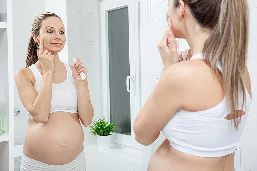Pregnant woman applying skincare in bathroom mirror.